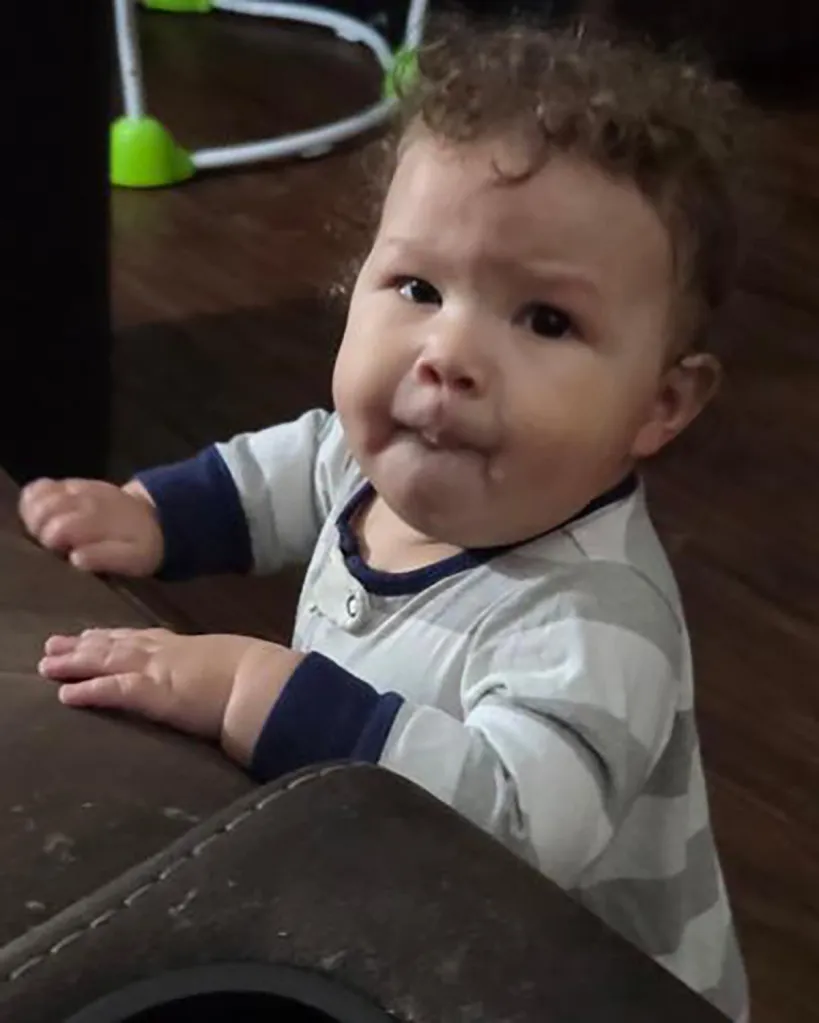 Remington Morningstar, a baby with curly hair, holding onto a dark brown piece of furniture.