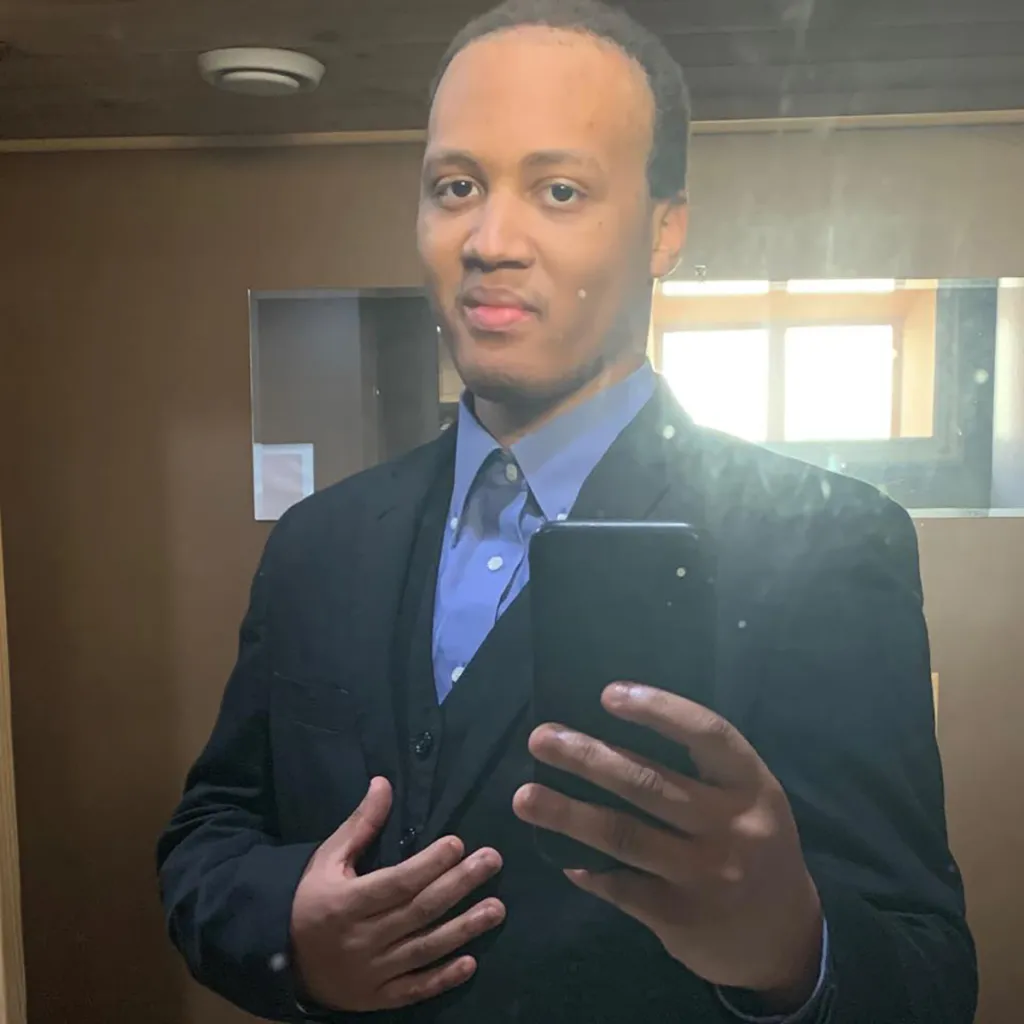 Omni Maxx Morningstar in a black suit and blue shirt, taking a mirror selfie.