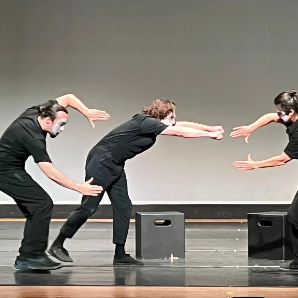 Three mimes in black clothing and white face paint performing on a stage with grey background.