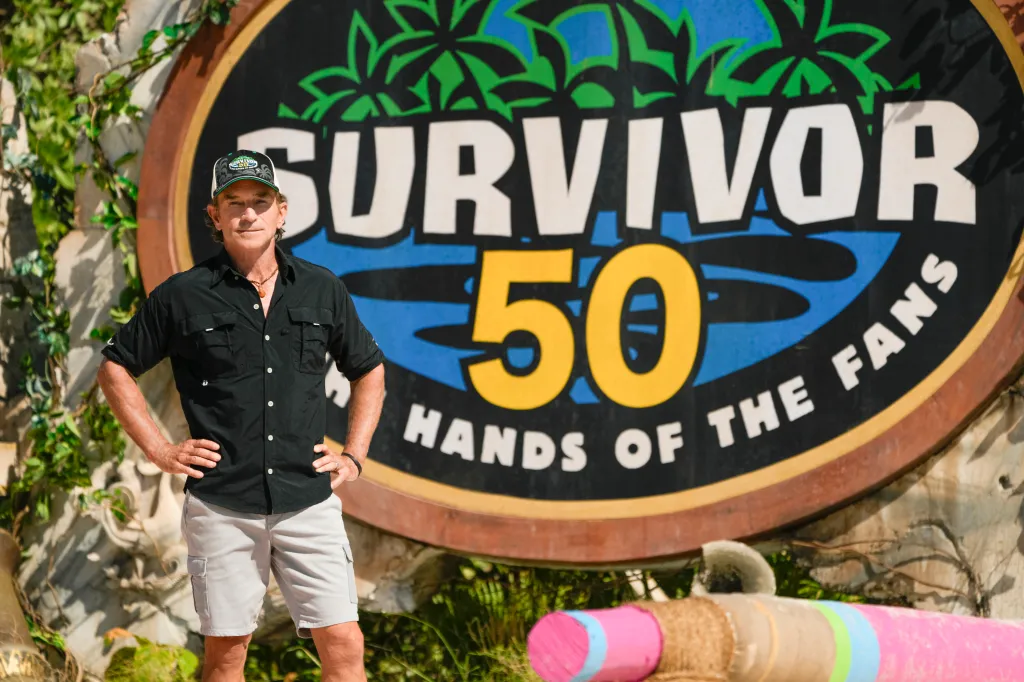 Jeff Probst stands in front of the Survivor 50: Hands of the Fans logo.