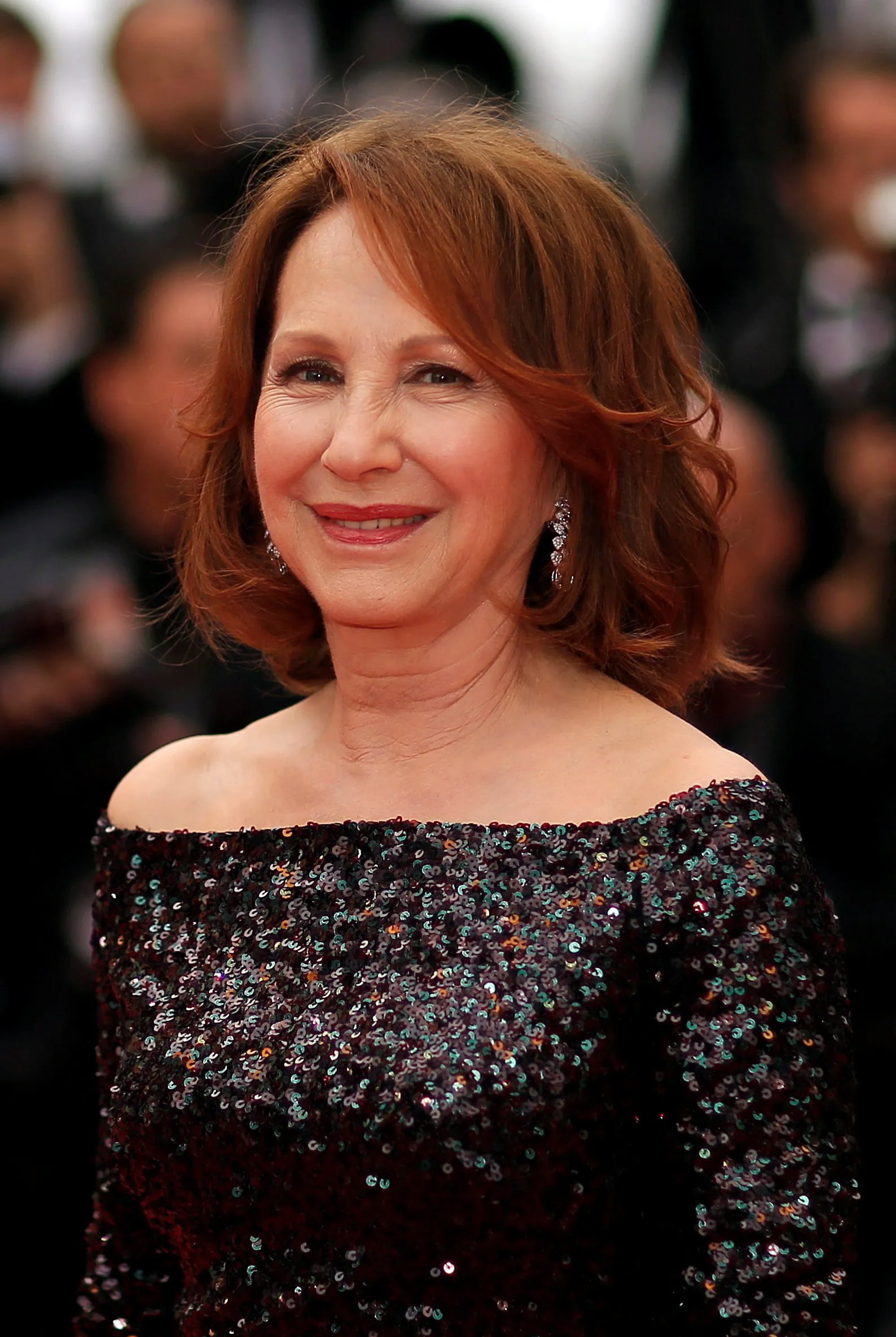 Nathalie Baye smiles at the camera, wearing a dark sequined off-the-shoulder dress.