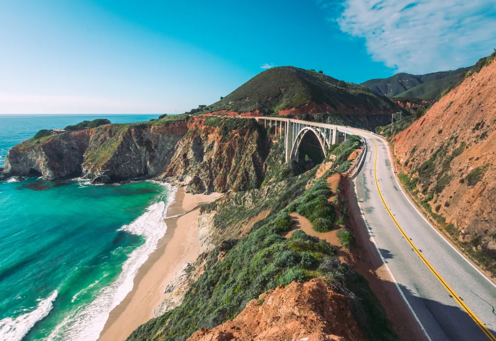 View of the Pacific coastline from Highway 1 in California, showing a bridge curving along green and red cliffs next to a sandy beach and turquoise water.