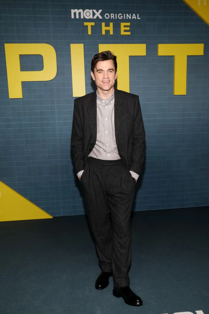 Patrick Ball poses in a pinstripe suit and light gray shirt, hands in pockets, in front of a blue grid background with yellow letters spelling 
