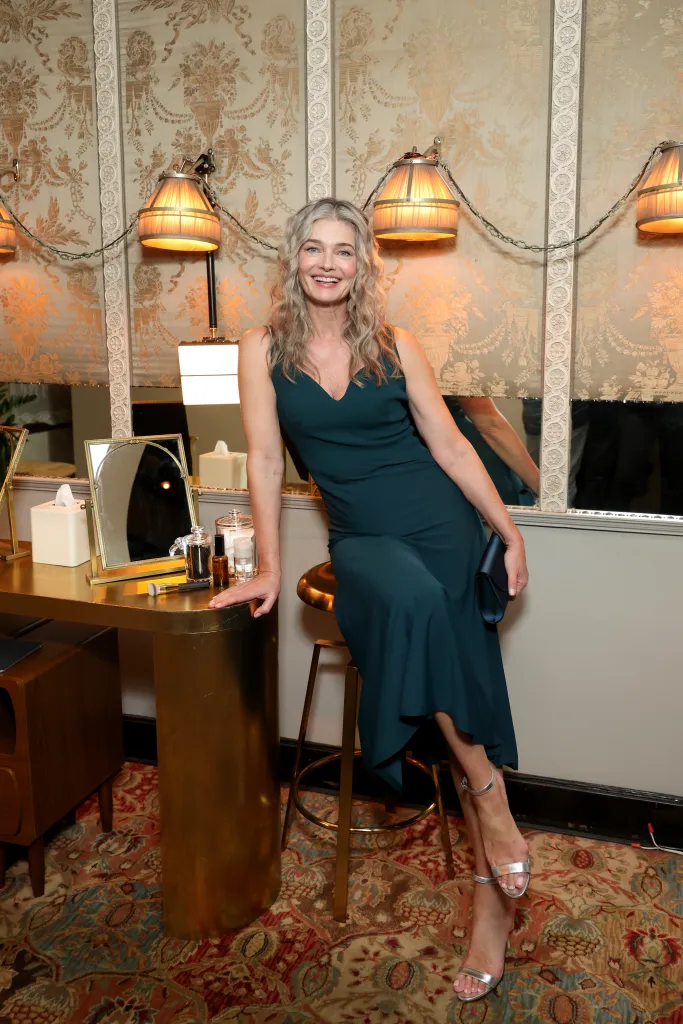Paulina Porizkova in a teal dress, seated on a stool next to a gold vanity, smiling at the camera.