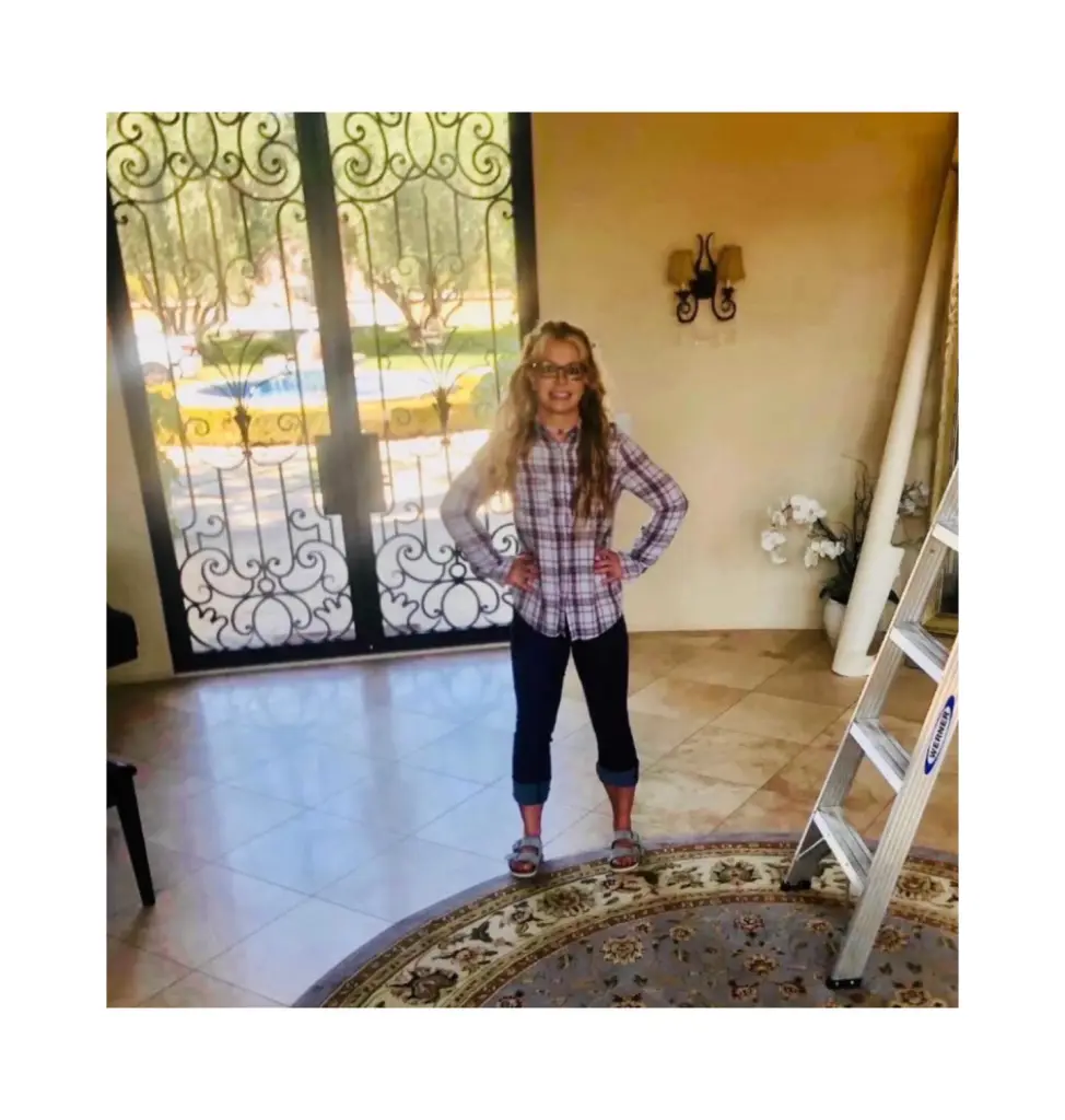 Britney Spears posing in a plaid shirt, dark pants, and sandals with hands on her hips, standing in a tiled room with ornate ironwork doors leading outside.