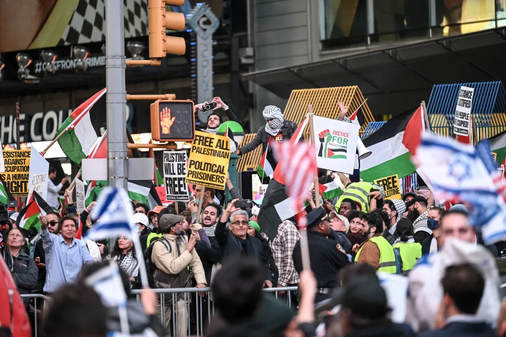 Pro-Palestine demonstrators wave Palestinian flags and hold signs at a rally in Times Square, New York, with some people in the foreground holding Israeli flags.