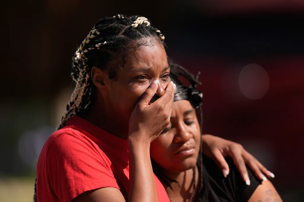 A woman with braids wipes tears from her face while another woman comforts her.
