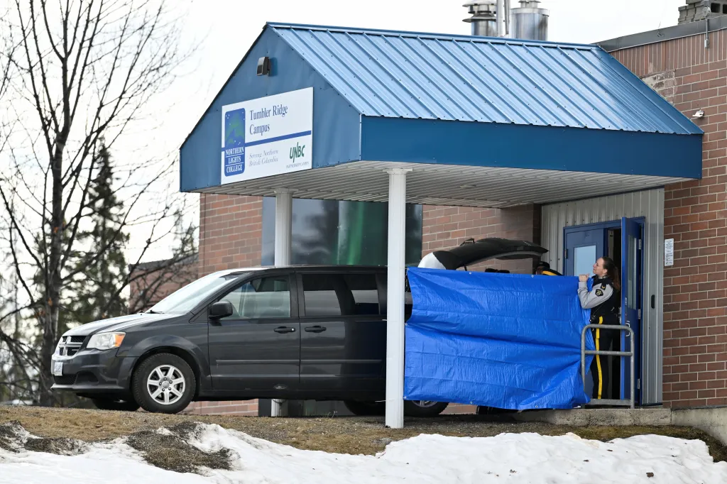 A police officer stands by the entrance of the Tumbler Ridge Campus building, partially covered by a blue tarp, with a dark minivan parked nearby.