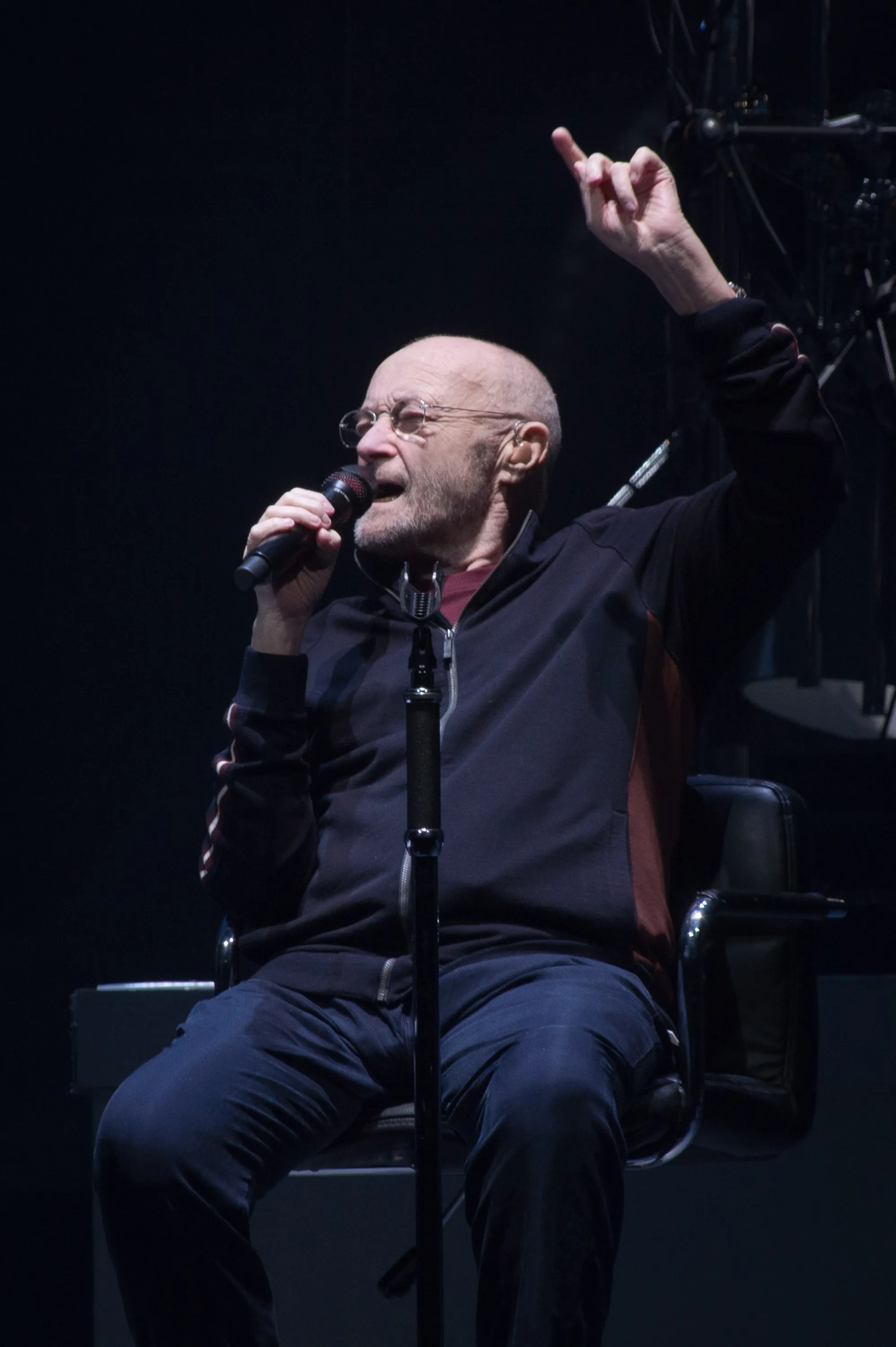 Phil Collins performing on stage, seated in a chair, holding a microphone, and raising his right arm.