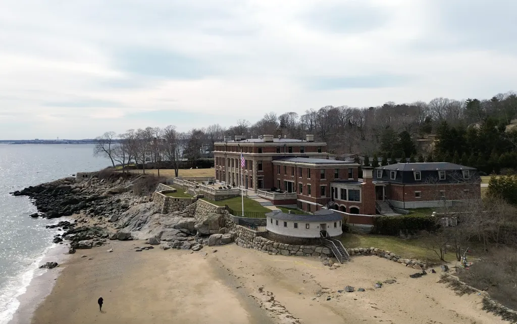 A drone view of a large brick waterfront mansion on a rocky coastline with a sandy beach and a person walking on it.