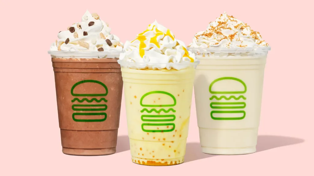 Three Shake Shack shakes in plastic cups, topped with whipped cream and various toppings.