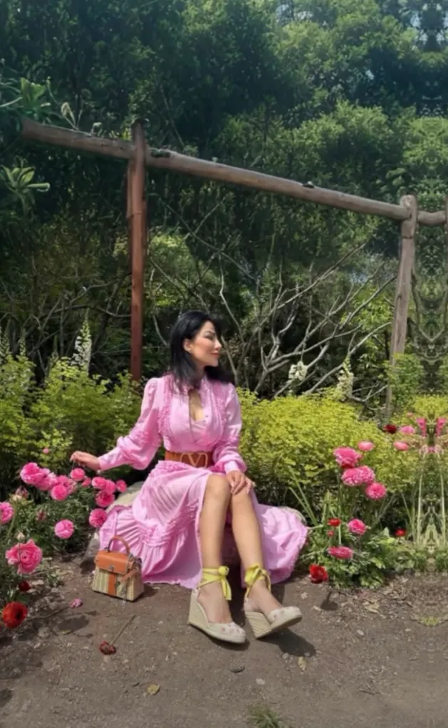 Hamideh Soleimani Afshar, niece of Qasem Soleimani, sits on the ground surrounded by pink flowers, wearing a pink dress, brown belt, and espadrille wedge shoes.