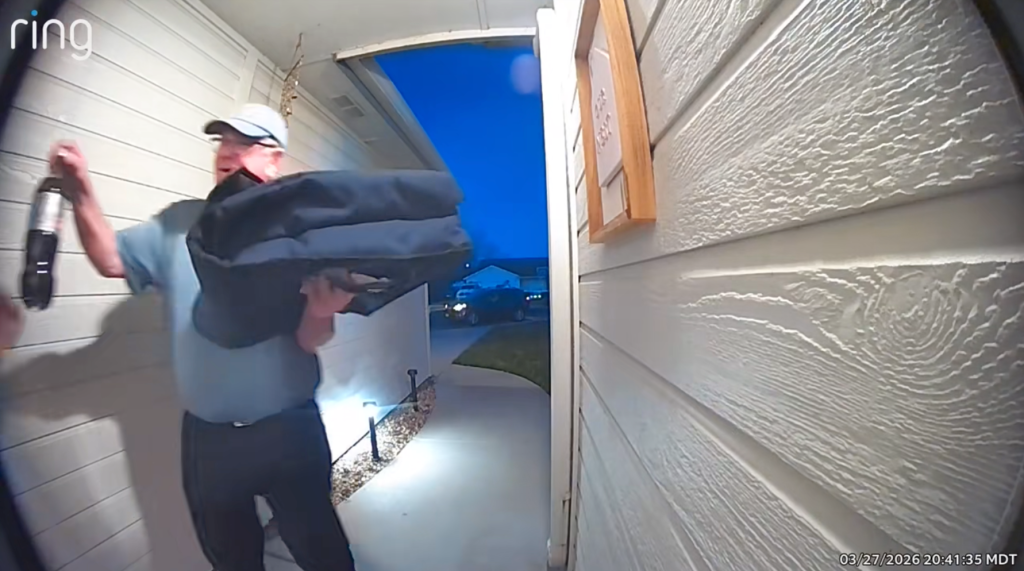 A person in a light blue polo shirt and hat holds a large black bag and a drink at a doorway.