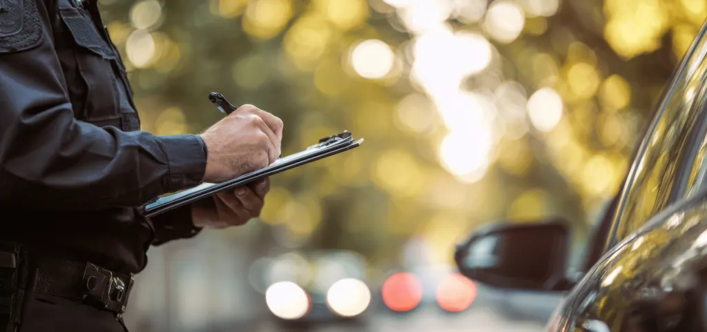 Police officer in uniform writing a parking ticket on a clipboard next to a car.