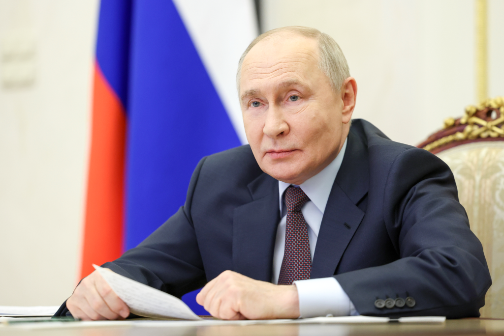 Russia's President Vladimir Putin sitting at a table with papers, with the Russian flag in the background.
