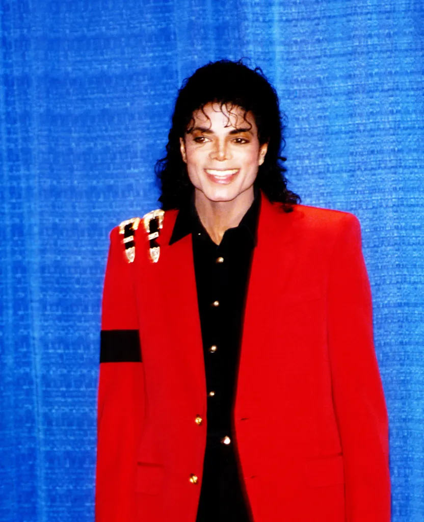 Pop singer Michael Jackson poses wearing a red sports coat with belt buckles on his right shoulder and a black arm band.