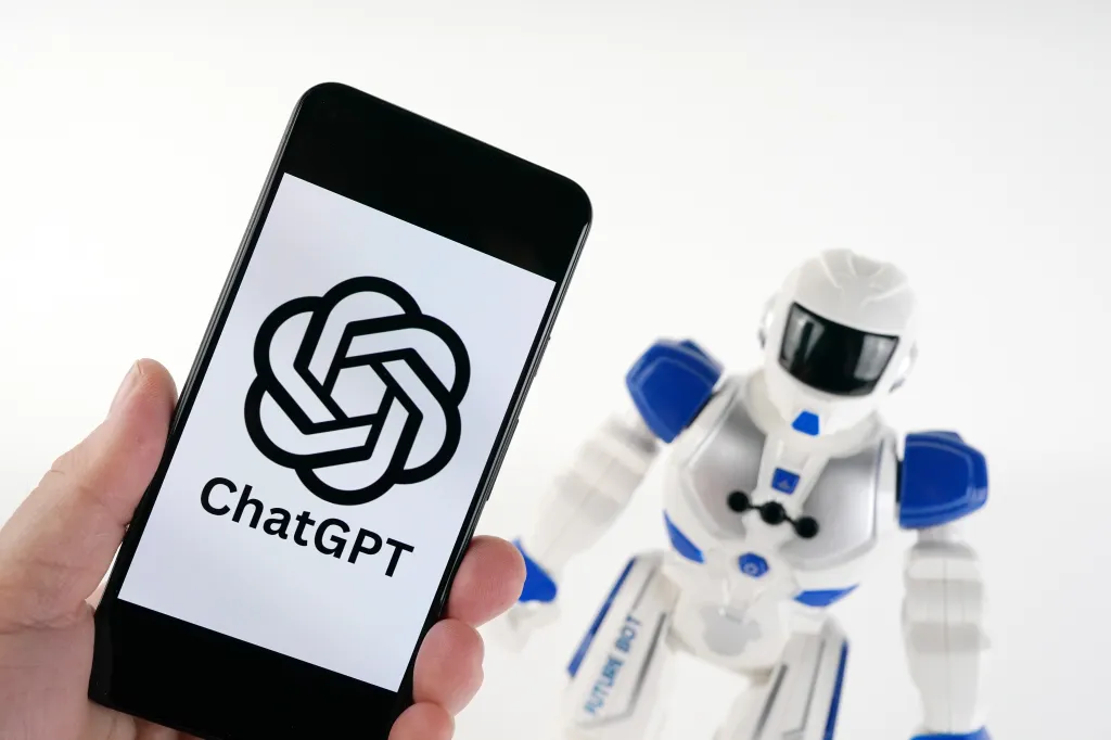 A hand holds a smartphone displaying the ChatGPT logo next to a white and blue toy robot.