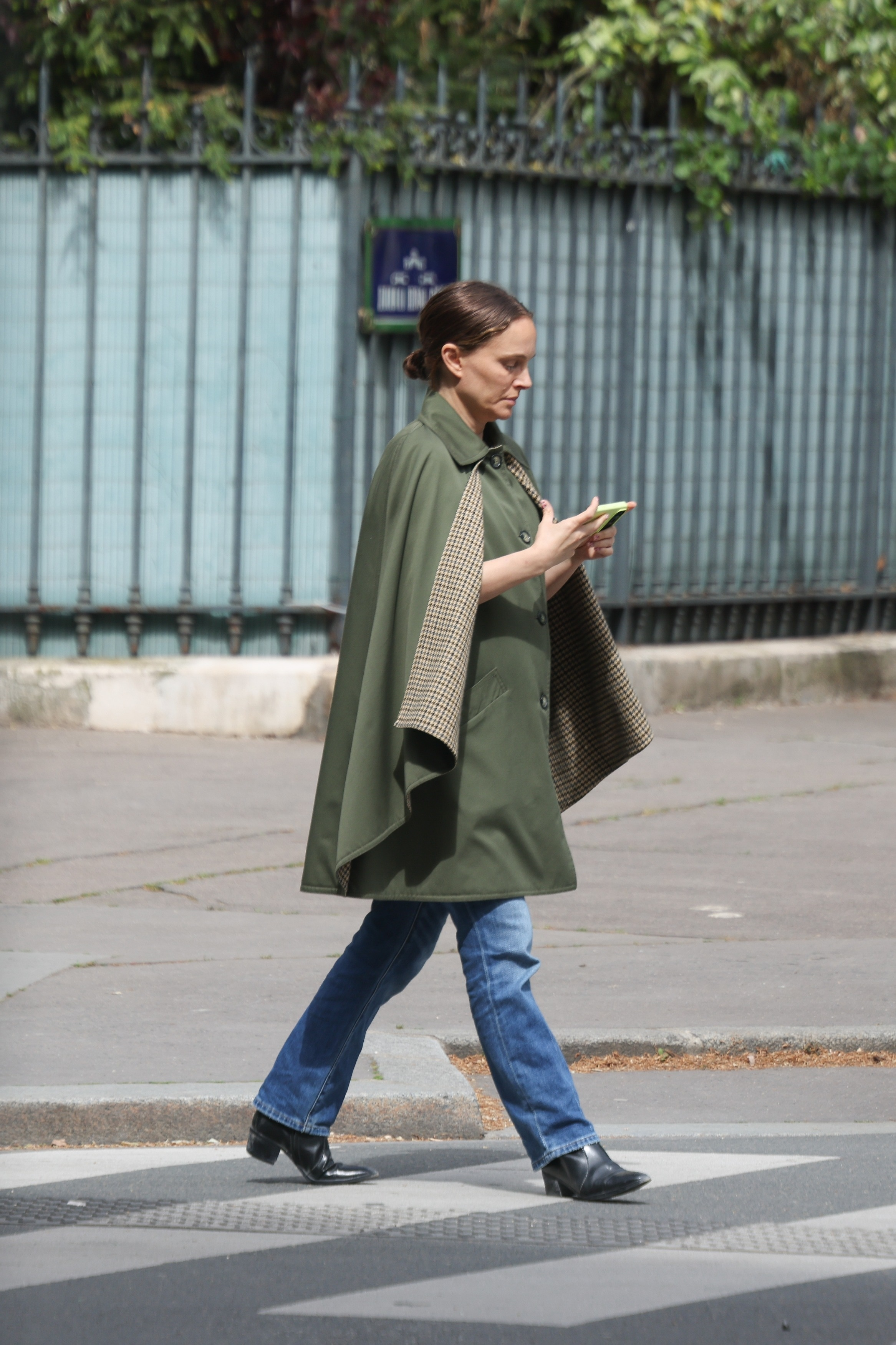 Natalie Portman wearing a green cape and jeans, looking at her phone while walking.