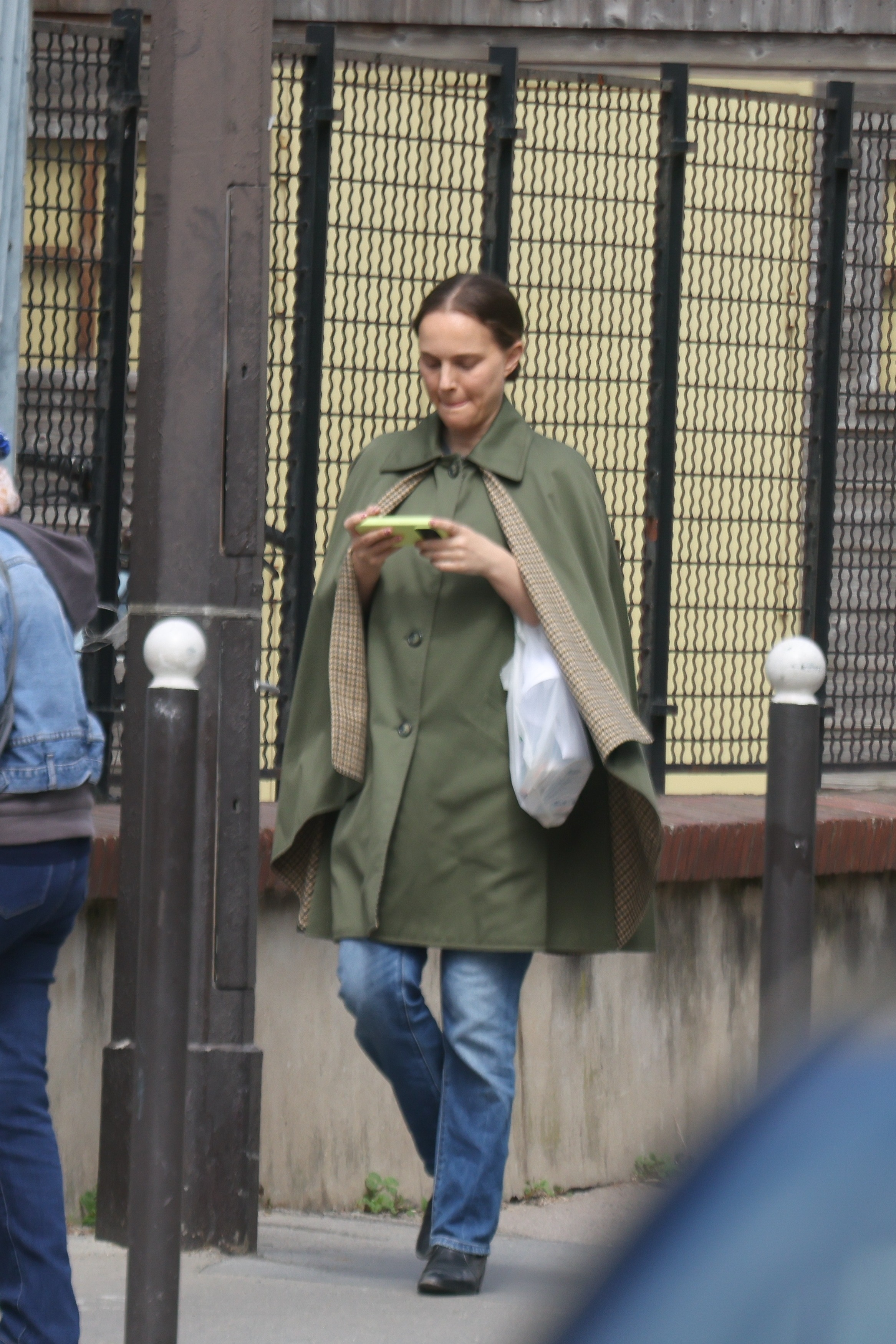 Natalie Portman wearing a green cape and blue jeans, looking at her phone.