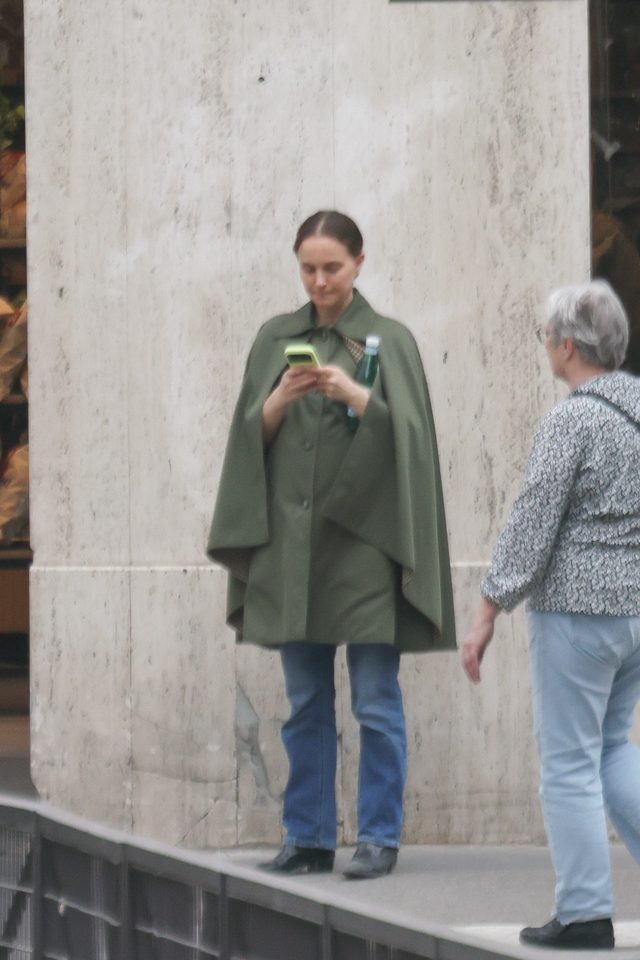 Natalie Portman looking at her phone while wearing a green coat and jeans.