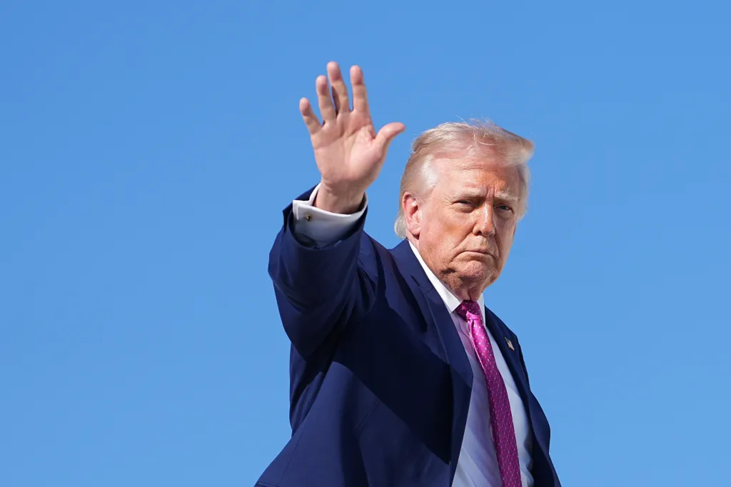 Donald Trump raises his right hand in a wave against a clear blue sky.