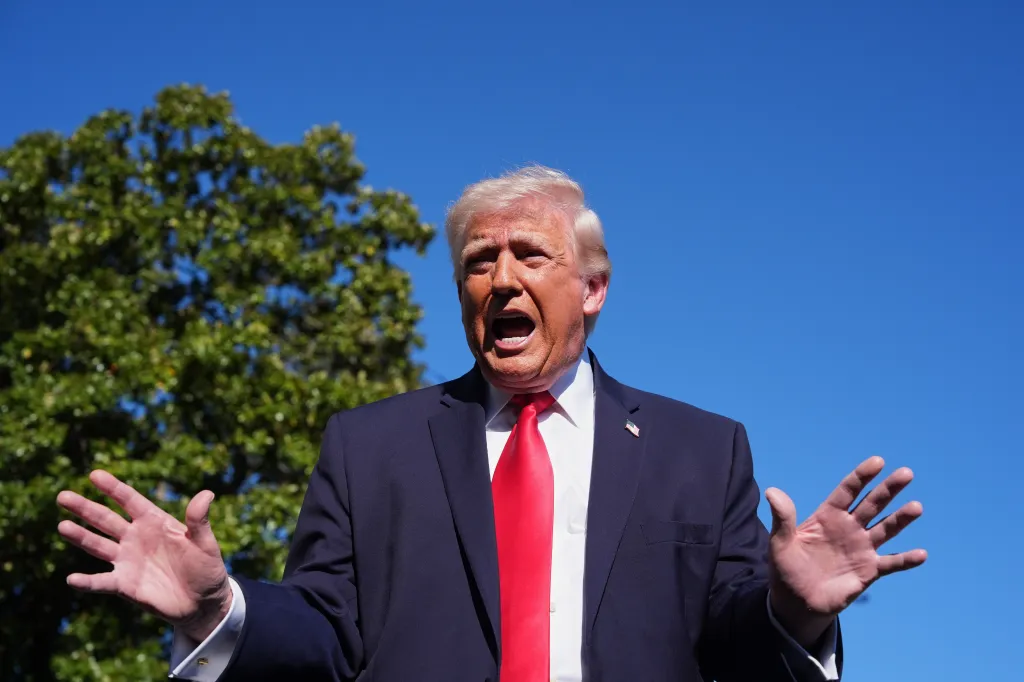 President Donald Trump speaking to reporters with his hands raised, wearing a suit and red tie, with a blue sky and green tree in the background.
