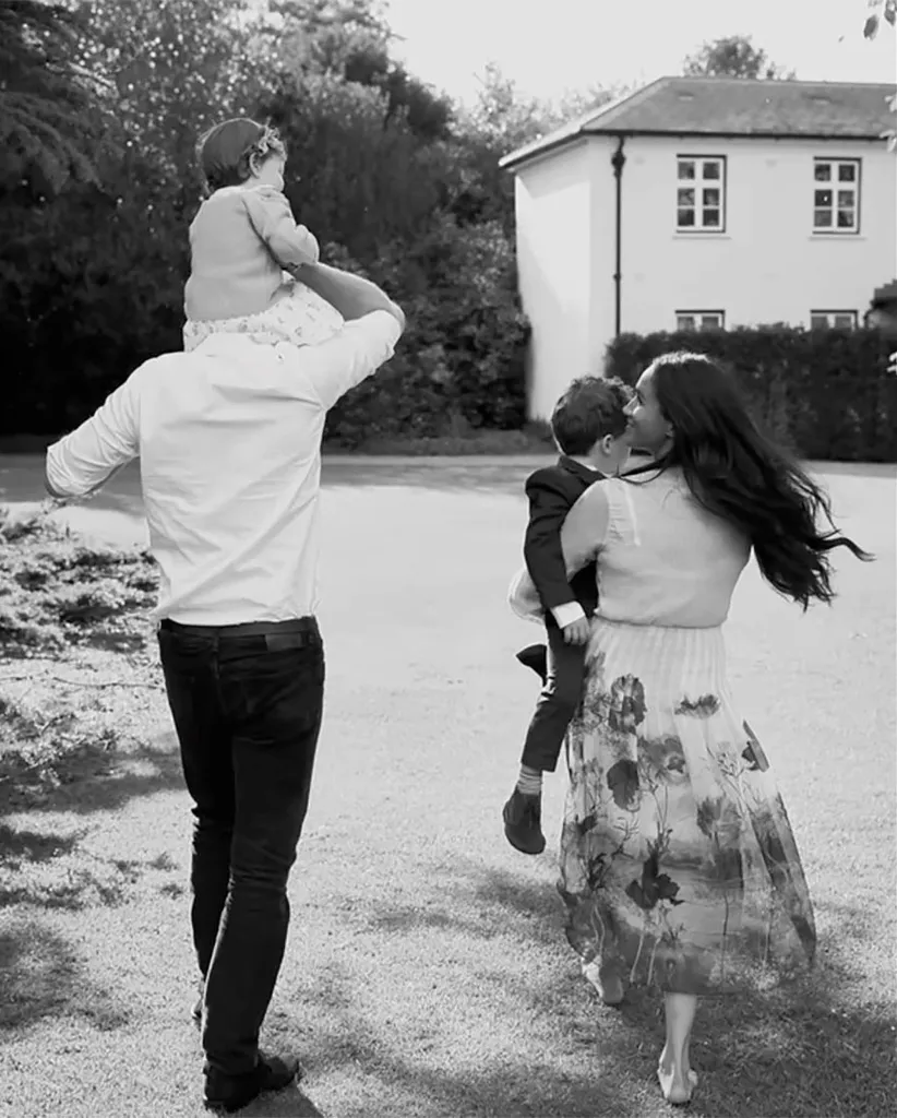 Black and white photo of a family of four walking away from the camera in a grassy area, a white house visible in the background.
