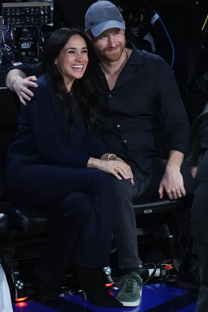 Prince Harry, in a gray baseball cap, with his arm around Meghan Markle, both smiling at an NBA game.