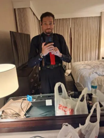 Man in a hotel room taking a mirror selfie, wearing a black shirt, red tie, and multiple knives and ammunition strapped to him.