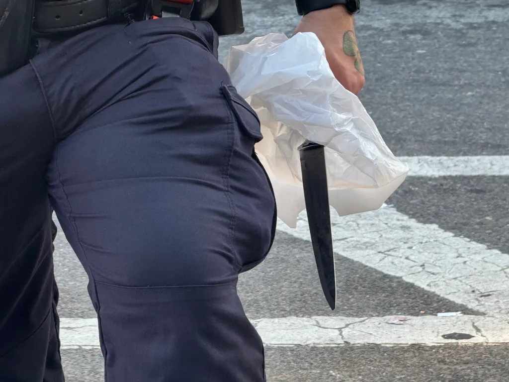 A large knife being held in a plastic bag by a person in blue uniform pants.