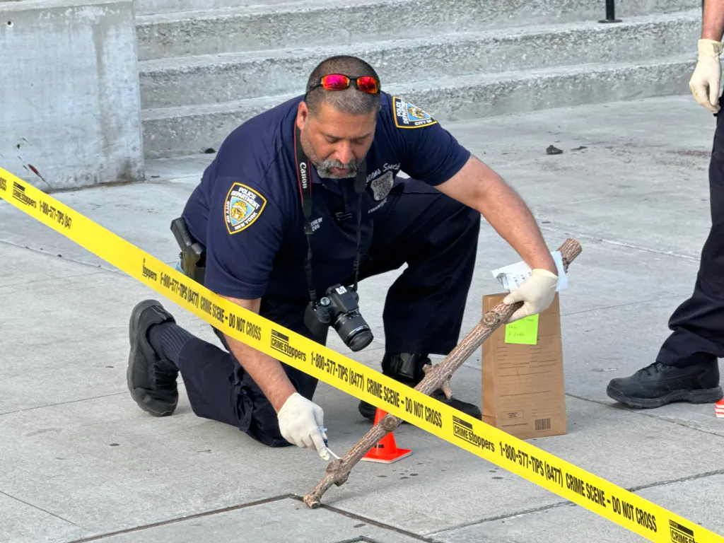 A police officer in gloves collects a large, gnarled stick from inside a roped-off crime scene.