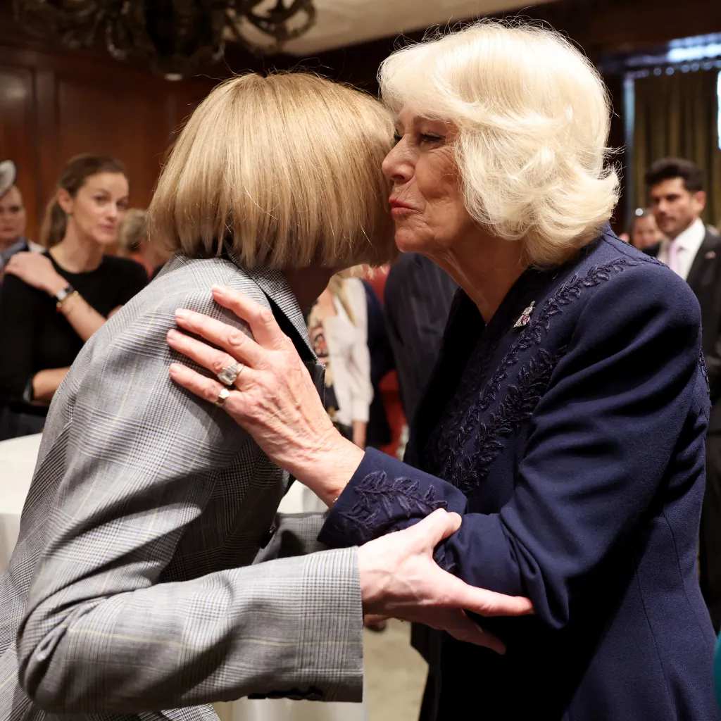 Queen Camilla, in a navy blue dress, embracing Anna Wintour, in a grey check skirt suit, at a literary event.
