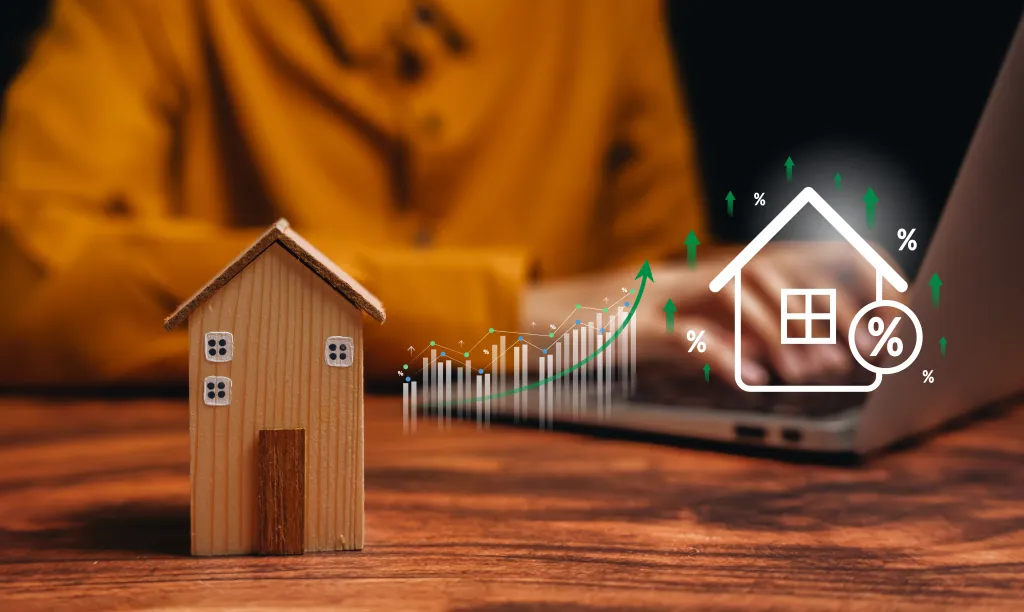 A wooden house model sits on a wooden table with a blurred background of a person working on a laptop, overlaid with a digital graph showing rising trends and a house icon with a percentage sign.
