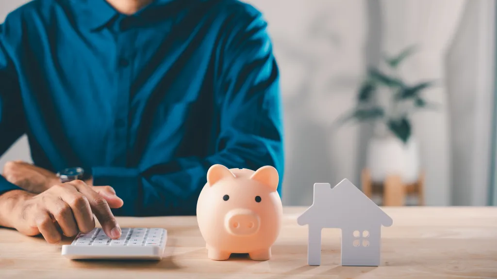 Man calculates finances for real estate investment with a piggy bank and house model on the table.