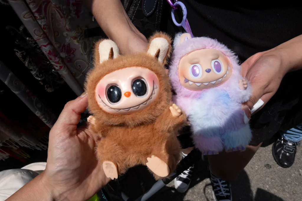 A person holding a brown Labubu toy, next to a multicolored Labubu toy being held by another person. The multicolored toy has a QR code on its tag.