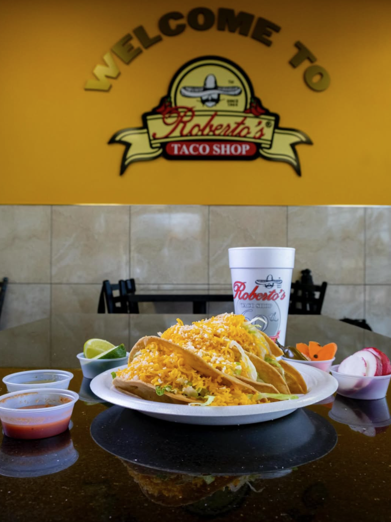 Three tacos on a plate with sides, a drink, and the Roberto's Taco Shop logo in the background.