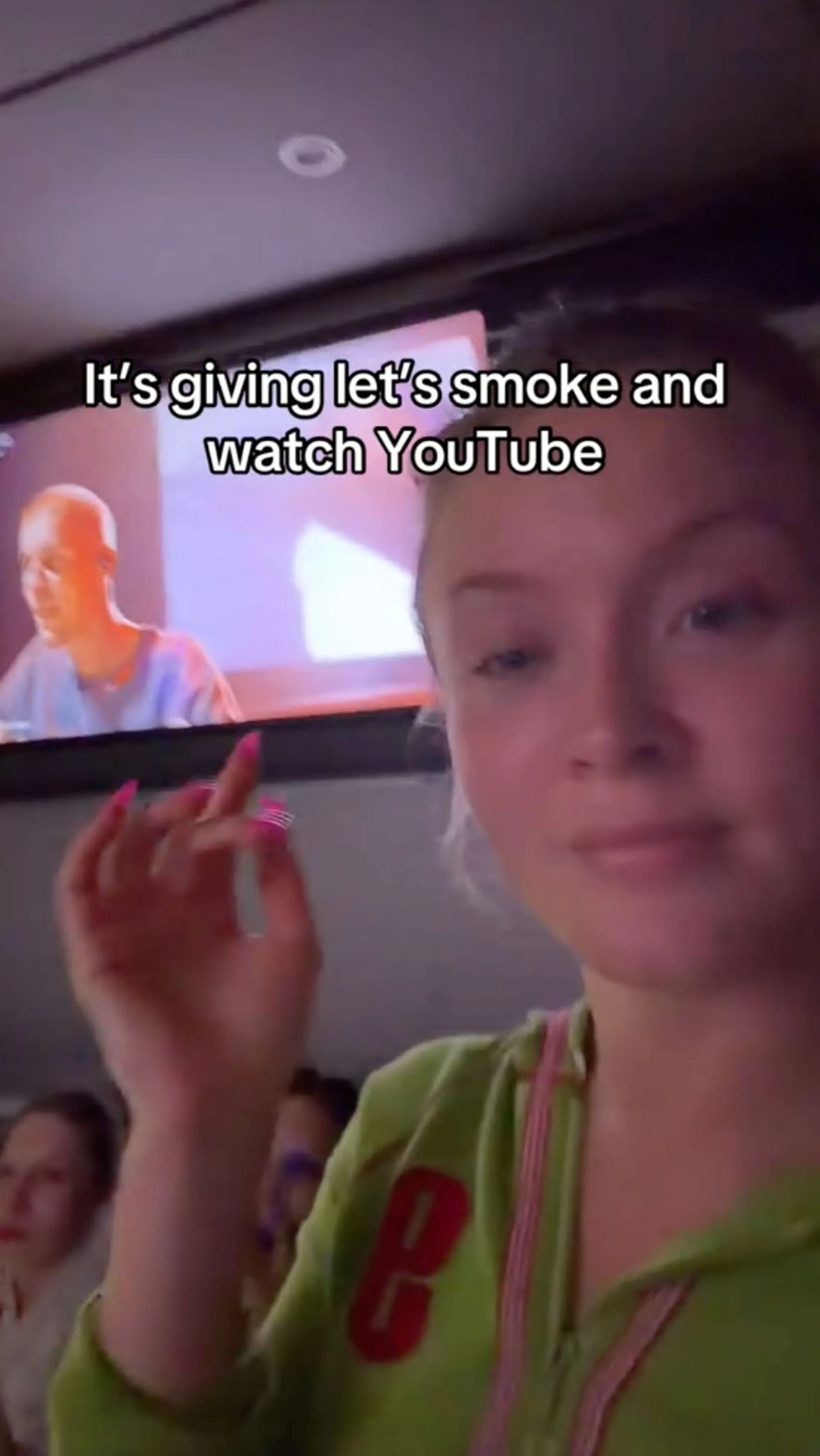 A woman in a green hoodie with pink drawstrings poses with a television screen behind her.