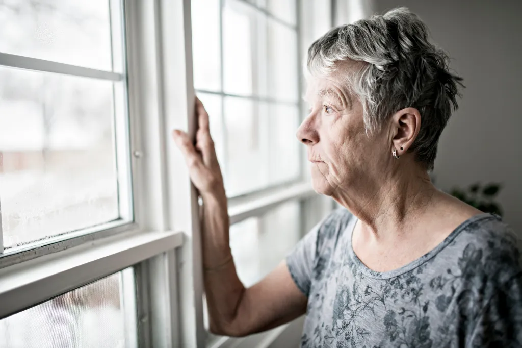 A sad, lonely elderly woman in her apartment looking out a window.