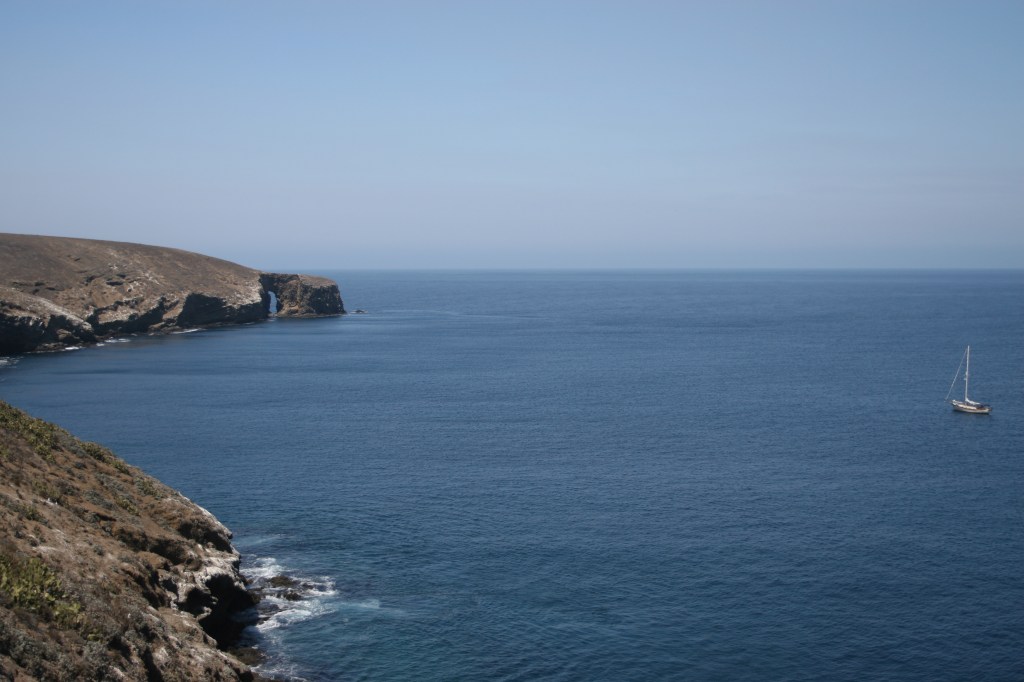 A lone sailboat anchored in the blue waters of Santa Barbara Island, California, with a rocky, desert-like coastline and a sea arch in the background.