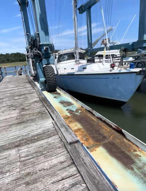 A blue sailboat docked beside a weathered wooden pier and boat hoist.