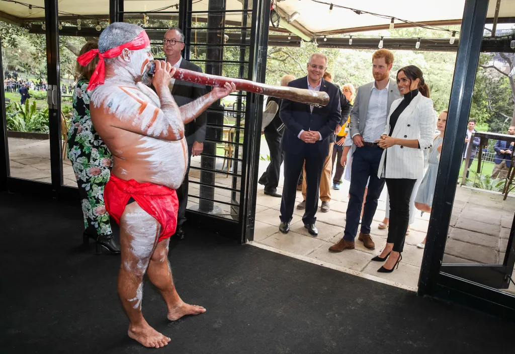 An Indigenous man in traditional paint and attire plays a didgeridoo for Prince Harry, in a grey blazer, white shirt and black pants, Meghan Markle, in a white blazer, black top and black pants, and Scott Morrison in 2018.