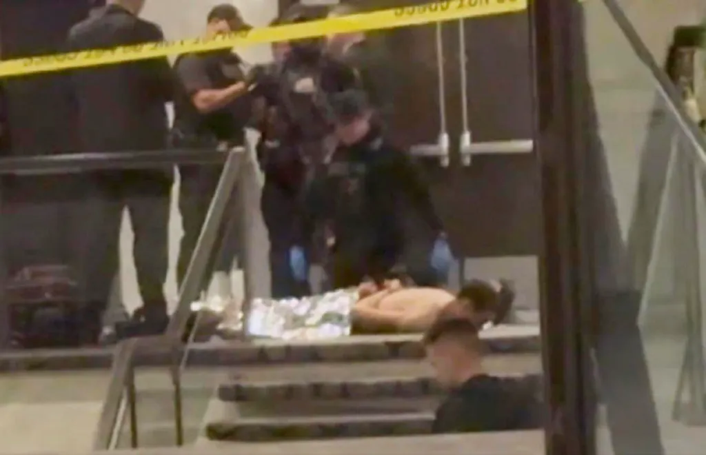 Law enforcement officers surround a shirtless individual lying on stairs under a yellow 