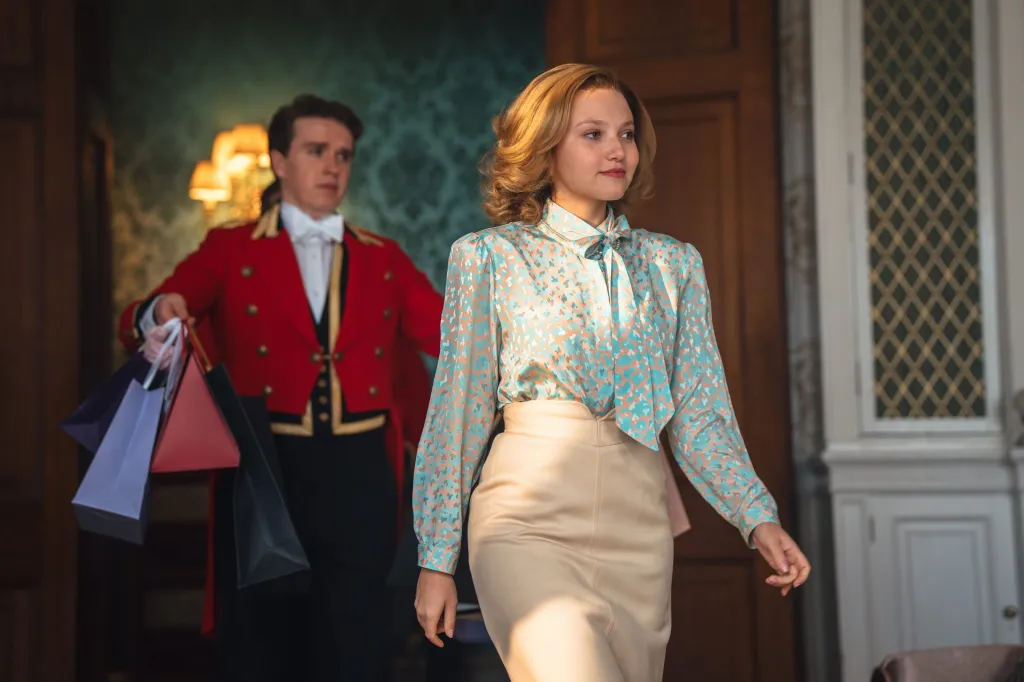 Mia McKenna-Bruce as Jane Andrews in a patterned blouse and cream skirt, followed by a man in a red uniform holding shopping bags.