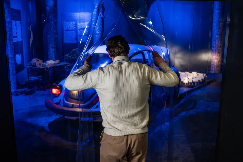 A man opening a clear plastic curtain to reveal a blue-lit exhibit with a white car in the background.
