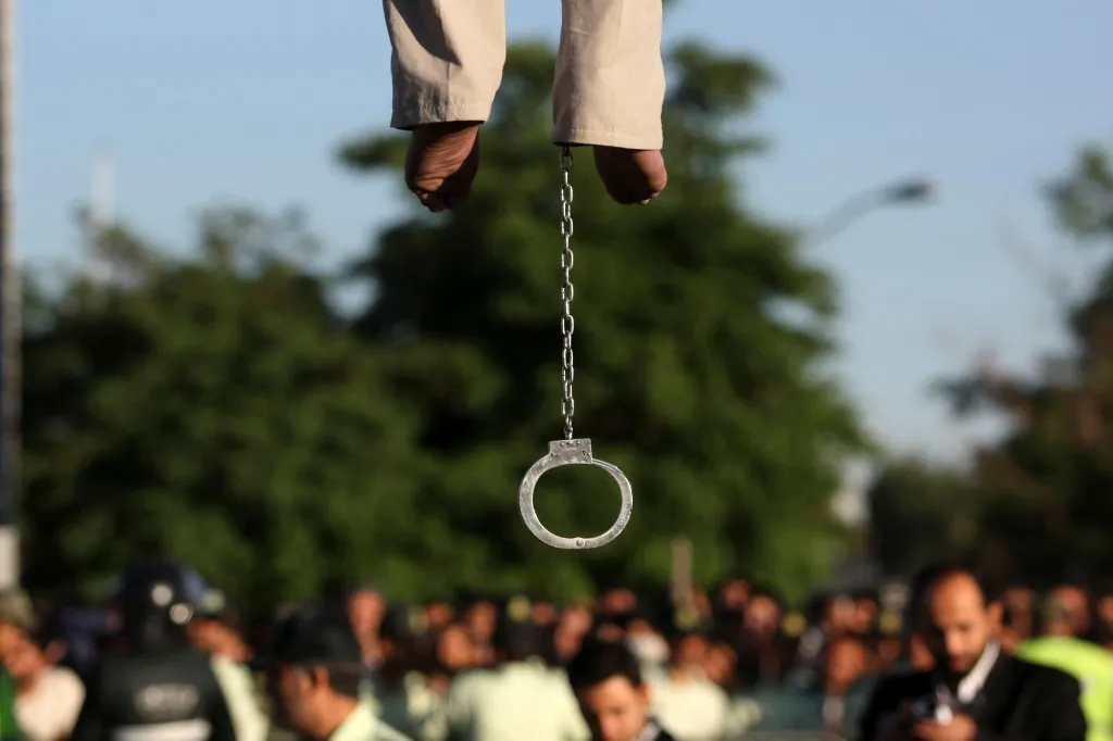 Feet of a publicly executed man with a shackle hanging below, as a crowd watches in the background.