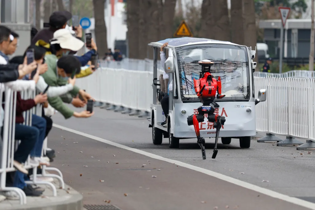 Shandian, a red humanoid robot, runs in a half marathon, escorted by a small white vehicle, while spectators film on their phones.