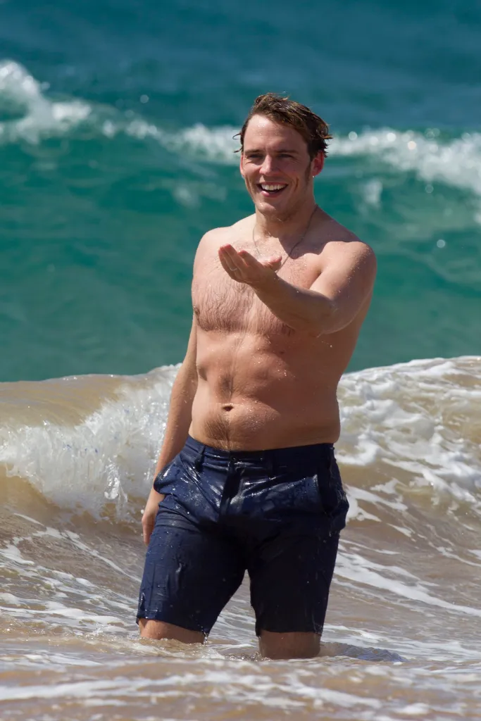 Sam Claflin smiling and extending his hand while in the ocean.