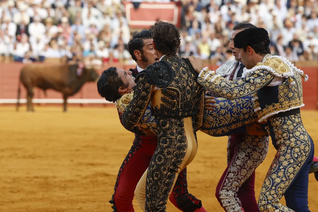 Bullfighter being carried by other bullfighters in an arena, with a bull in the background.