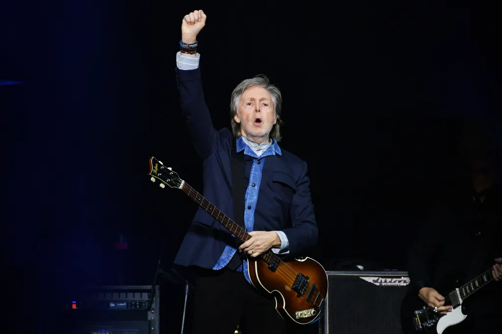 Paul McCartney performing on stage, holding a bass guitar and raising his left fist.