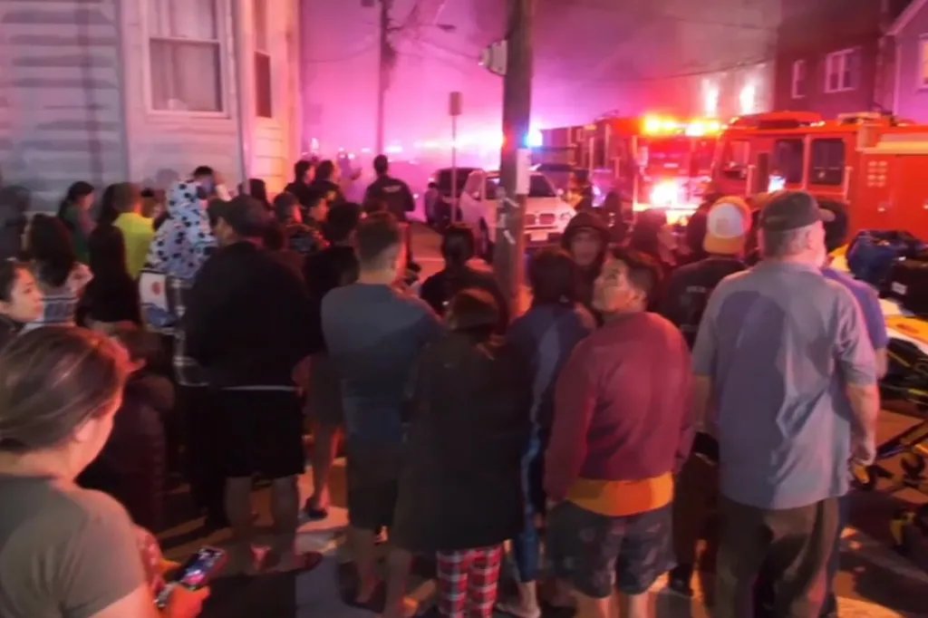 Crowd of people looking towards a building engulfed in a fire with emergency vehicles.